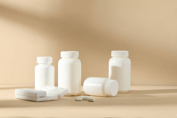 A group of white medicine bottles without label featured on beige background, next to some white tablets flat lay and a white pill box. Front view, copy space for text